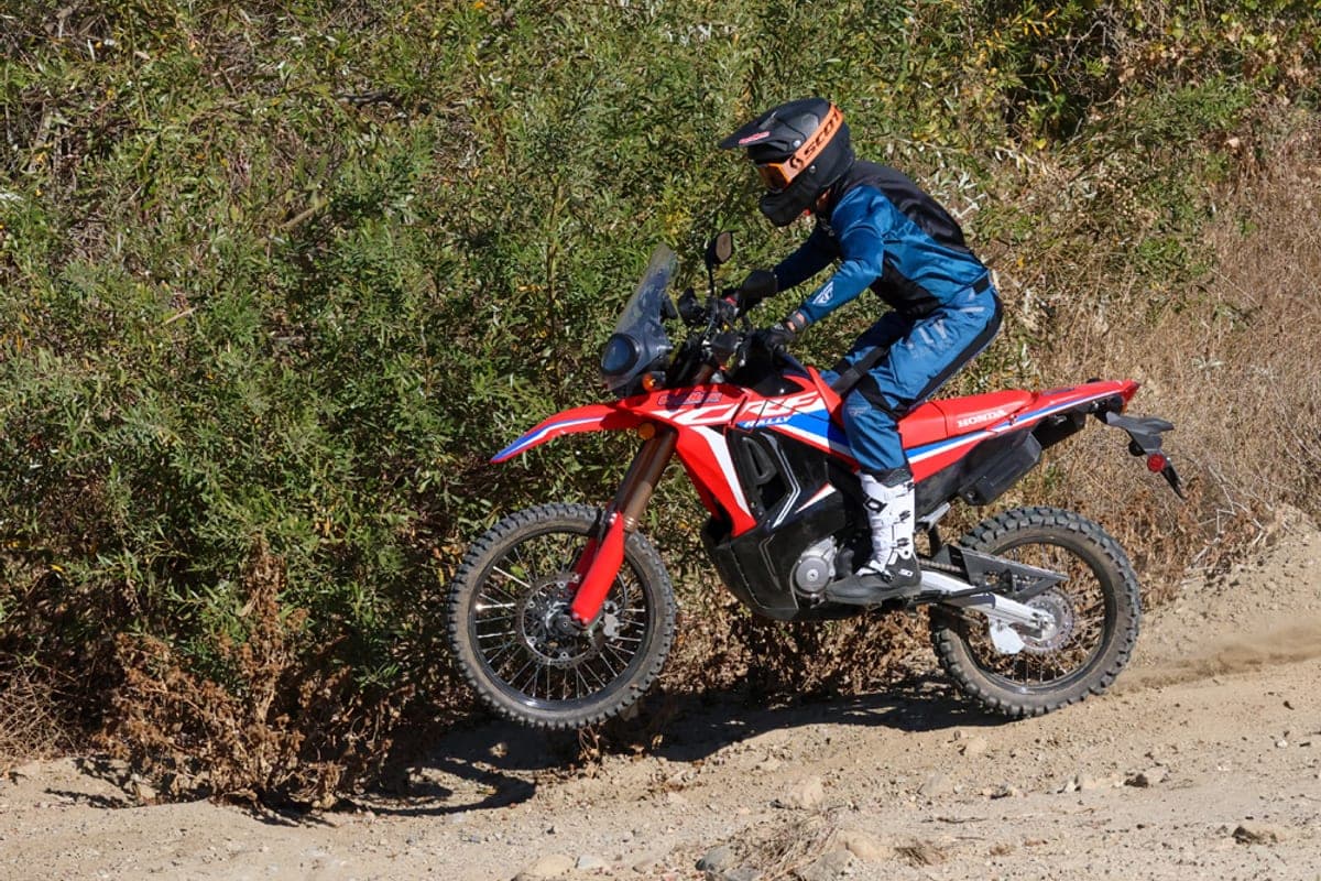 Rider in blue riding gear and a helmet powering the red Honda CRF300L Rally up a dusty trail, kicking up a dirt roost from the rear tire