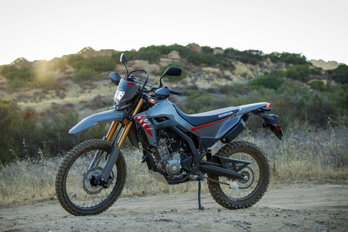 2026 Honda CRF300LS parked on a dirt trail at dusk, three-quarter view with scrubland hills and a dirt road in the background