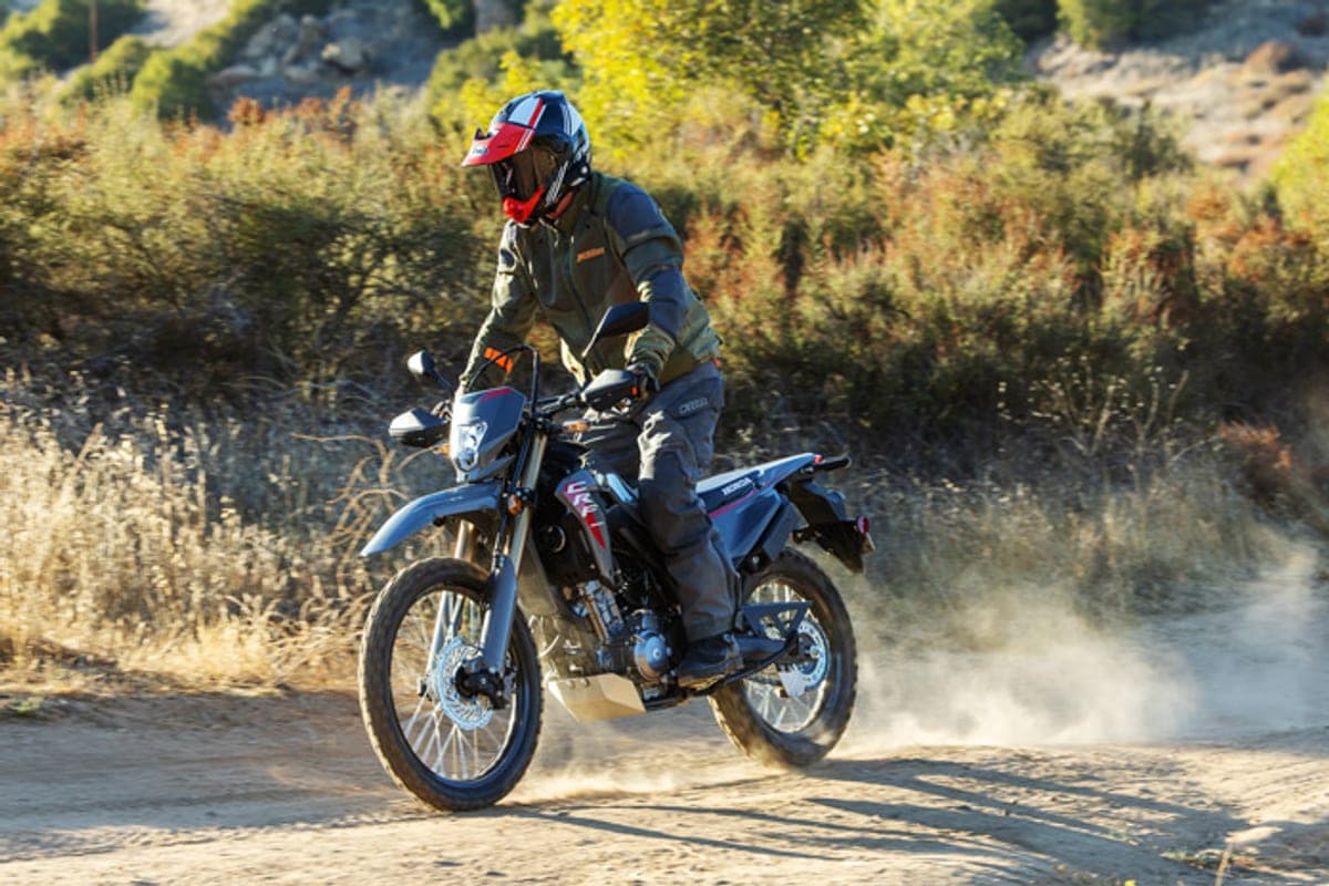 Rider in olive gear and a red-white-blue helmet powering a Honda CRF300LS through a dusty turn on a sandy trail