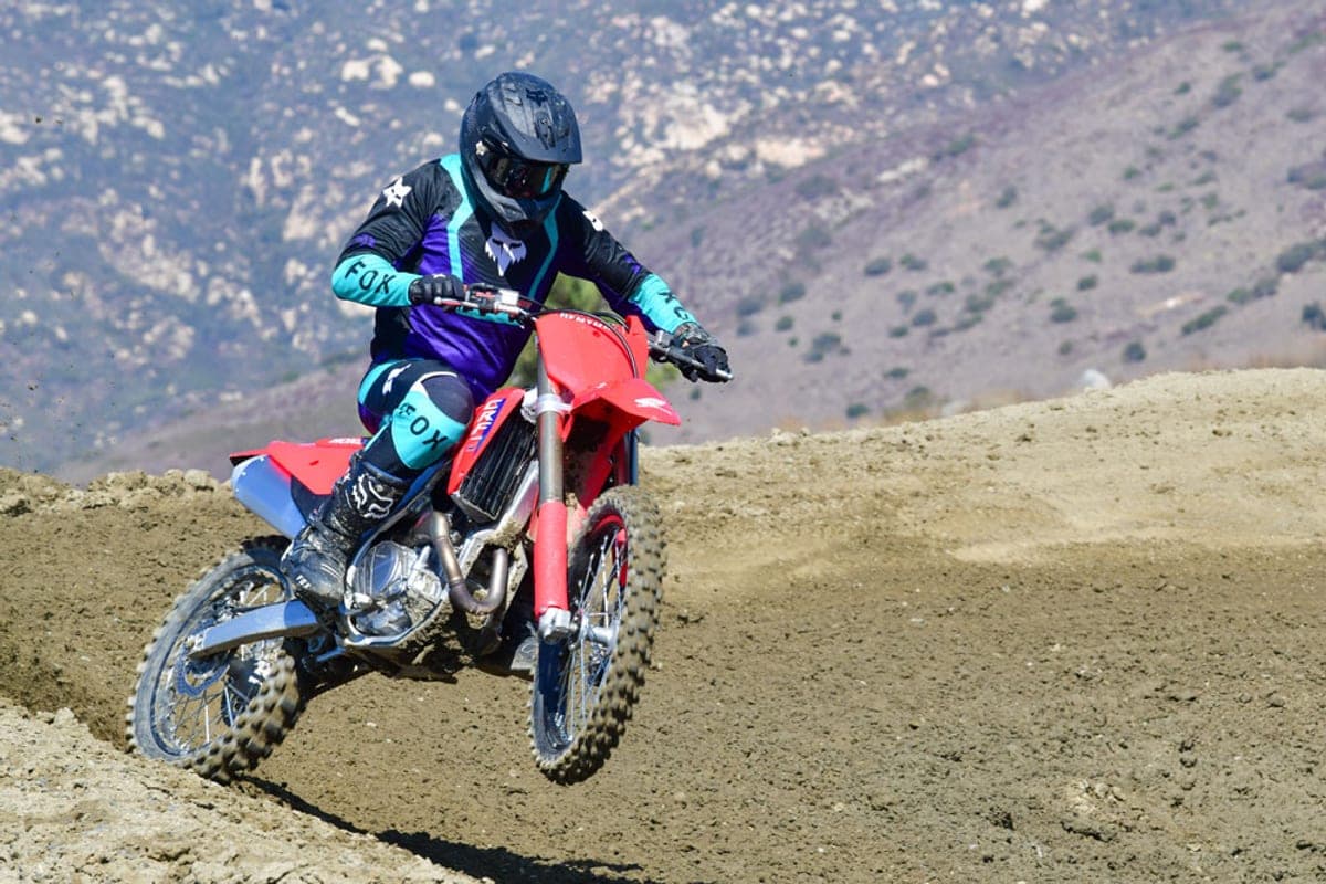 Rider in black and teal Fox gear launching a red Honda CRF450R off a dirt jump on a motocross track with hazy mountains behind