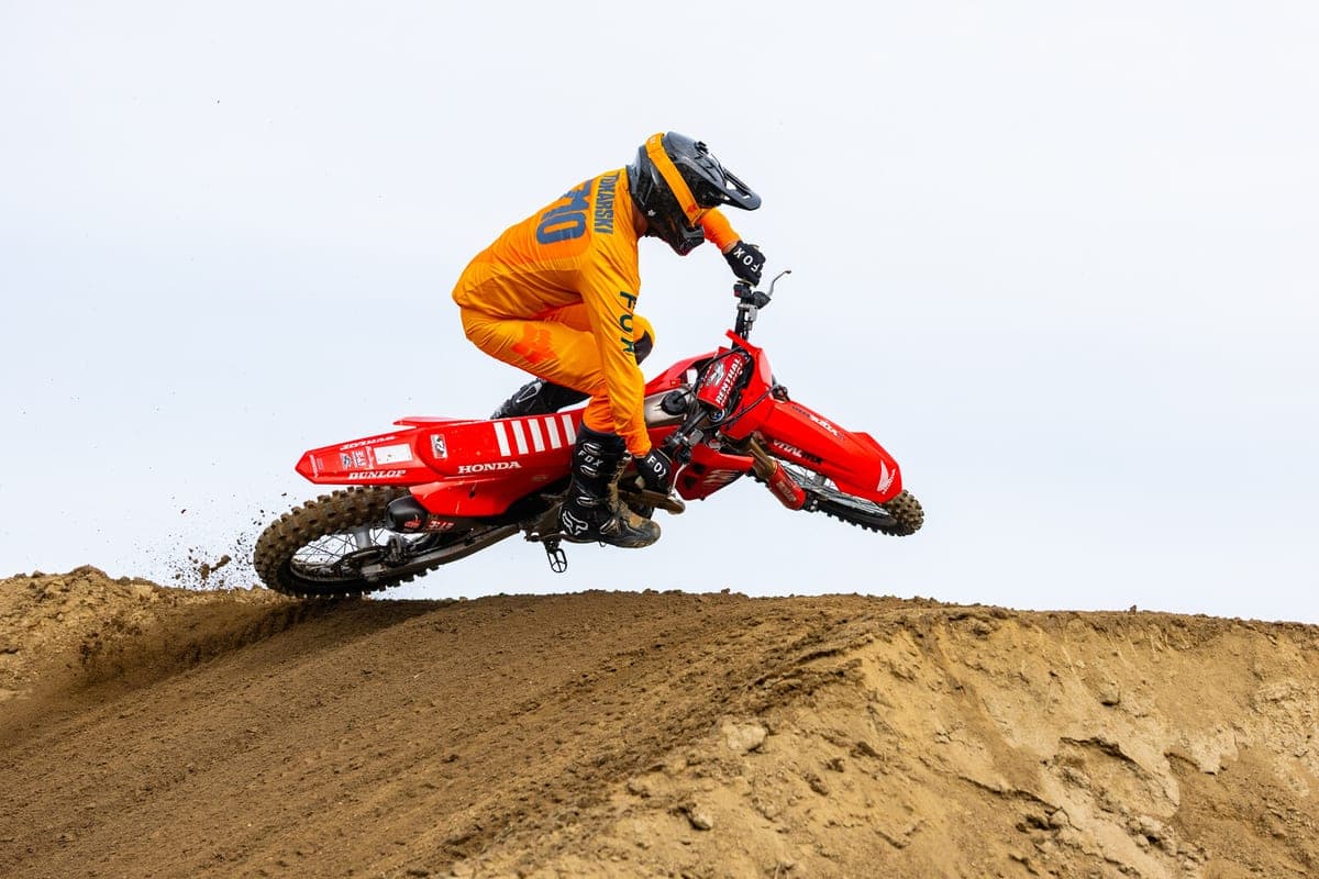 Rider in orange gear whipping a red Honda CRF450R HRC off a motocross jump, kicking up a spray of tan dirt