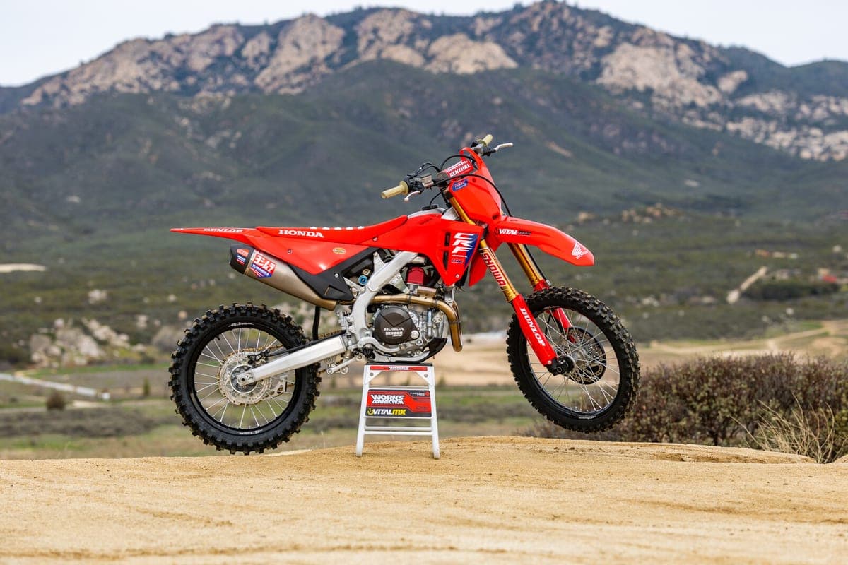 Honda CRF450R HRC Works Edition parked on a stand at a dirt track, left-side profile with hills and mountains in the background