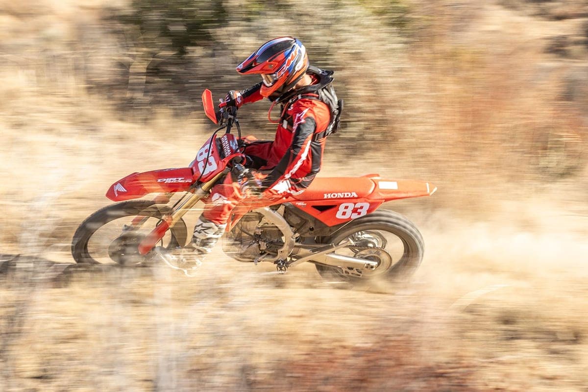 Rider in red and black gear leaning into a fast turn on a red Honda CRF450RX, kicking up a heavy cloud of dust on a dry trail