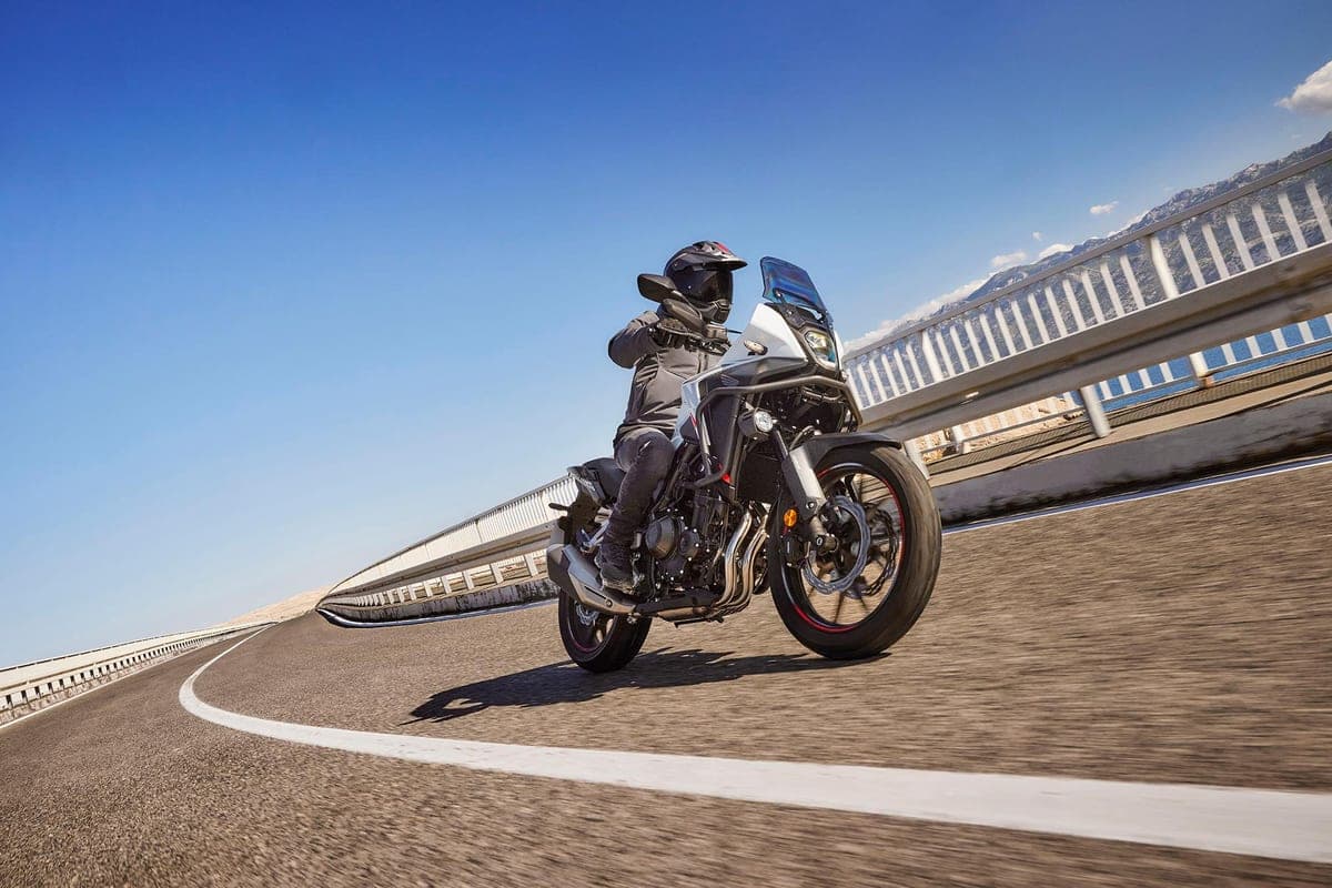 Rider in dark gear leaning the white Honda NX500 through a mountain road curve, front wheel lofting slightly with a guardrail and hazy peaks behind