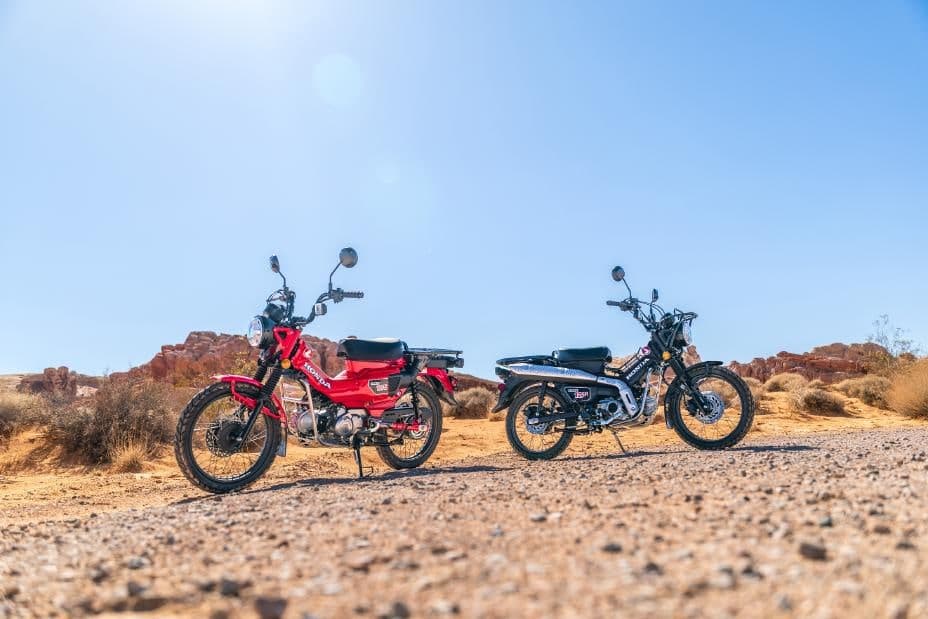 Red and black Honda Trail125 models parked side by side on a gravel desert trail with red rock formations under a clear sky