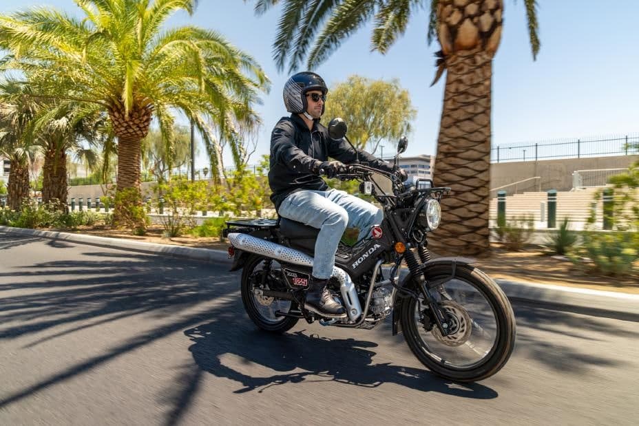 Rider in a black jacket and open-face helmet cruising a black Honda Trail125 past palm trees on a paved street