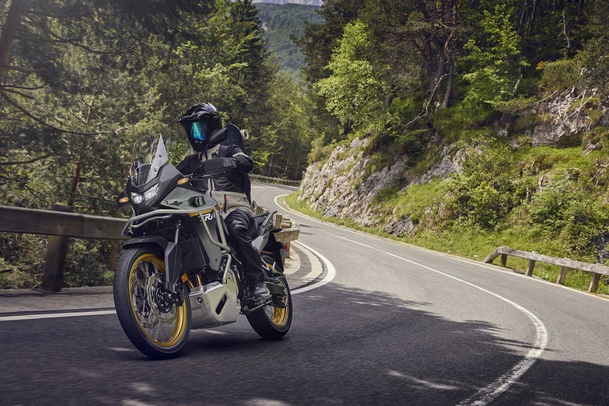 Rider in black gear on a grey Transalp XL750 leaning through a forested mountain road curve, gold wheels and crash bars visible