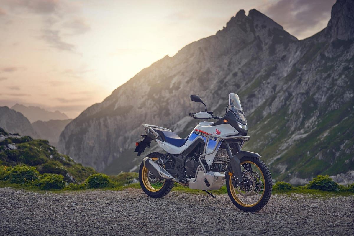 White and blue Honda Transalp XL750 parked on a gravel overlook at sunset, rear three-quarter view with rugged mountain peaks behind it