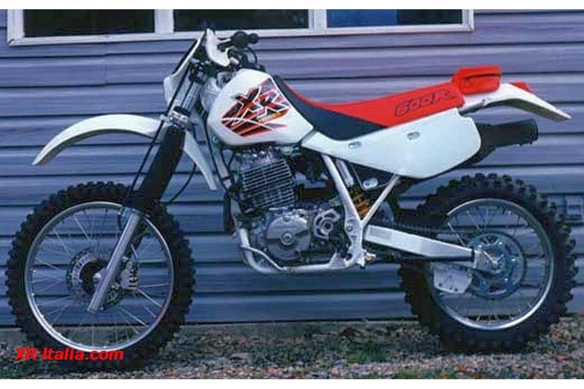 Honda XR600R in white bodywork with red seat and tank graphics, left-side view parked against a house's gray siding, showing the radiator and skid plate