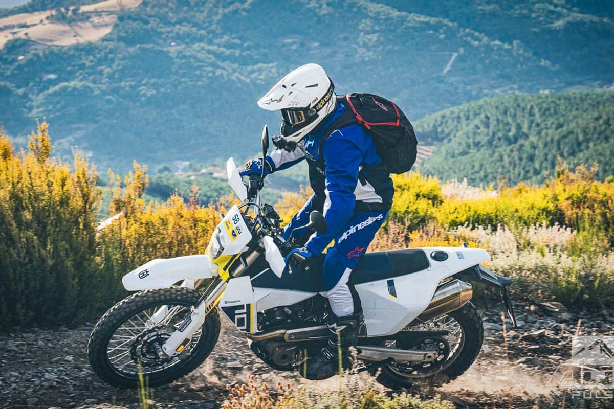 Rider in blue Alpinestars gear and white helmet leaning into a rocky hillside trail on the white and yellow 701 Enduro, kicking up dust
