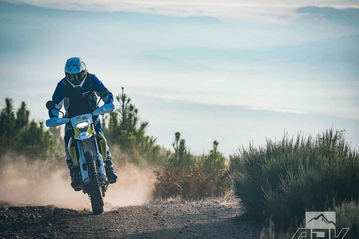 Husqvarna 701 Enduro headlight mask view as the rider powers up a dusty dirt track with pine-covered hills in the background