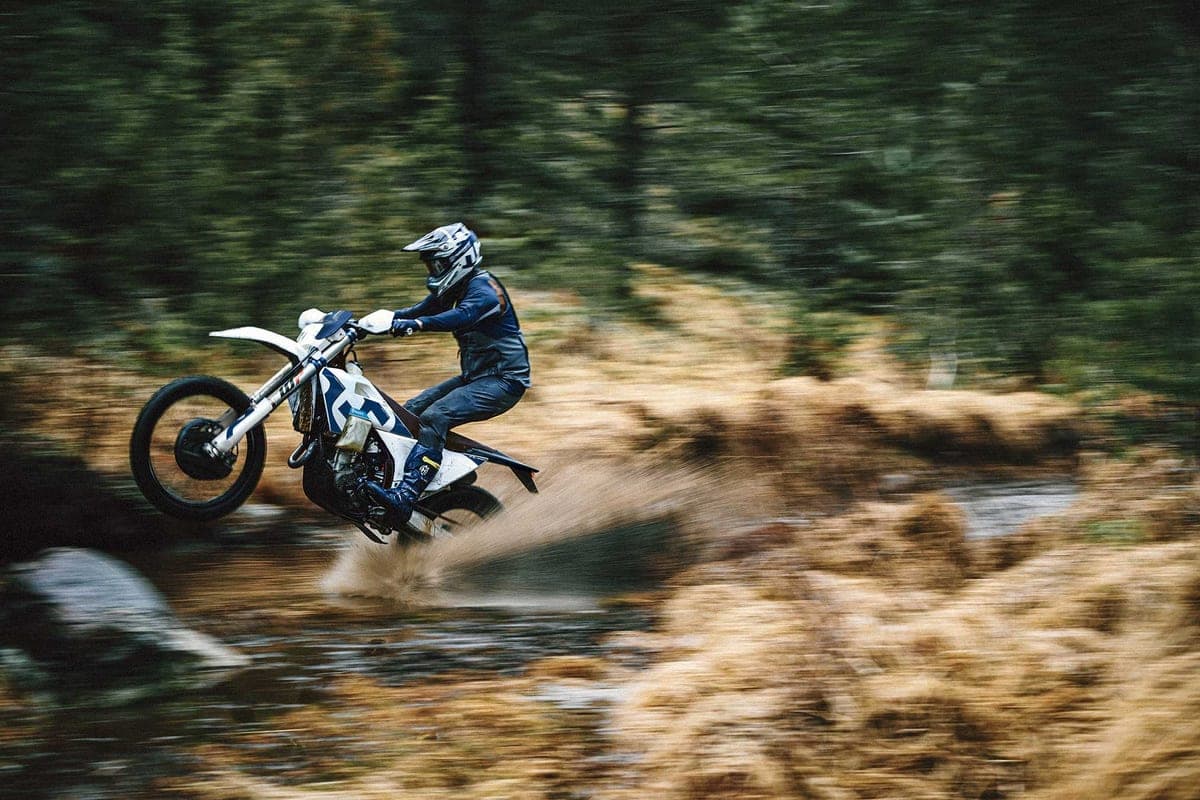 Rider in dark gear powering a Husqvarna FE 350s through a shallow creek crossing on a forest trail, kicking up a spray of water