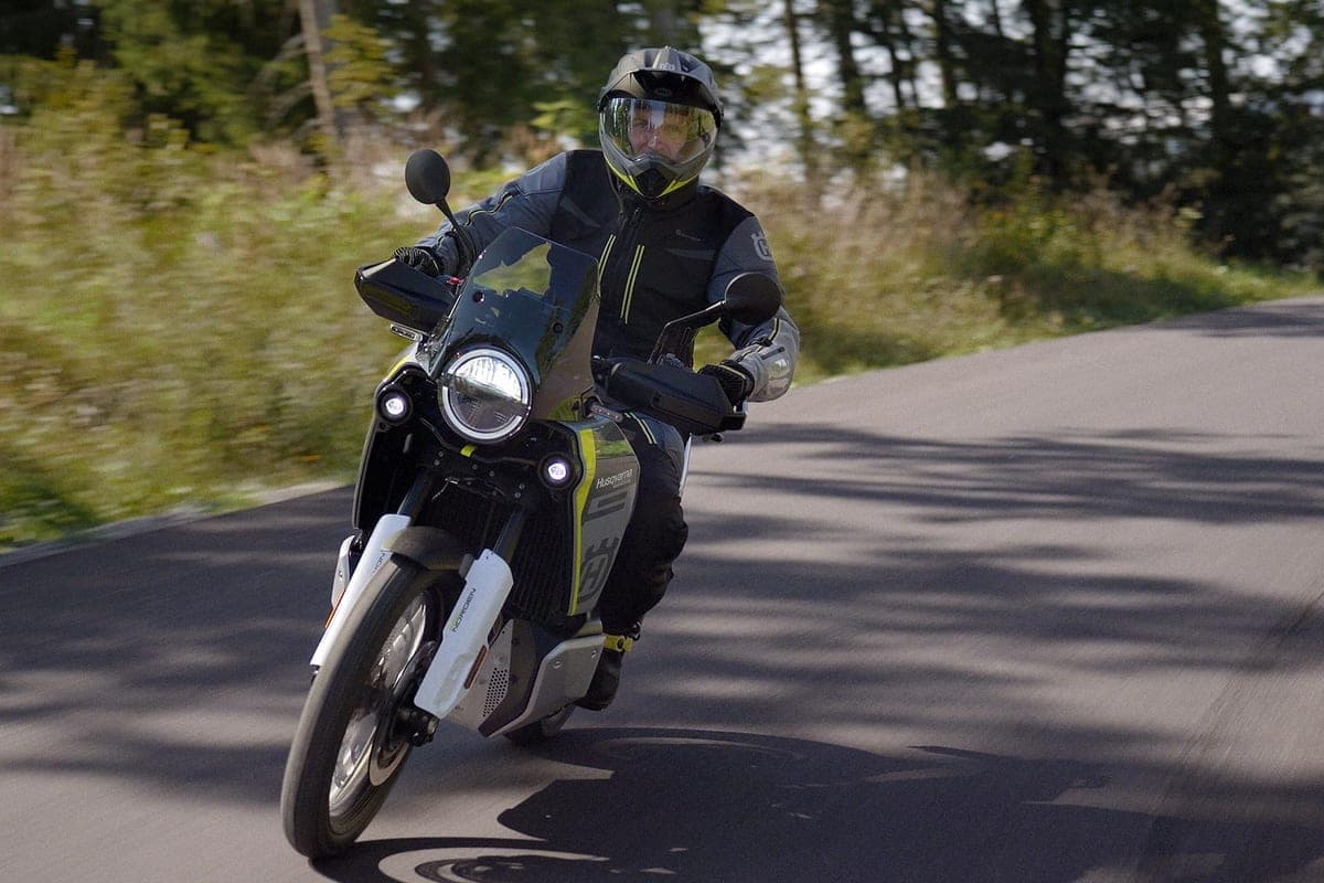 Rider in black and yellow riding gear leaning into a corner on a paved forest road, round LED headlight lit and windscreen deflecting the air