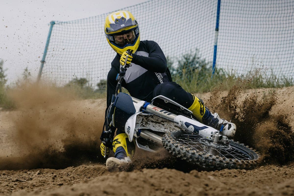 Rider in yellow gear leaning hard into a sandy motocross berm on a Husqvarna TC 125, kicking up a wall of dirt spray