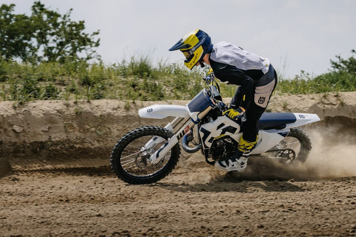 Husqvarna TC 125 rider carving through loose dirt in white and navy gear, front wheel cutting a groove as dust trails behind