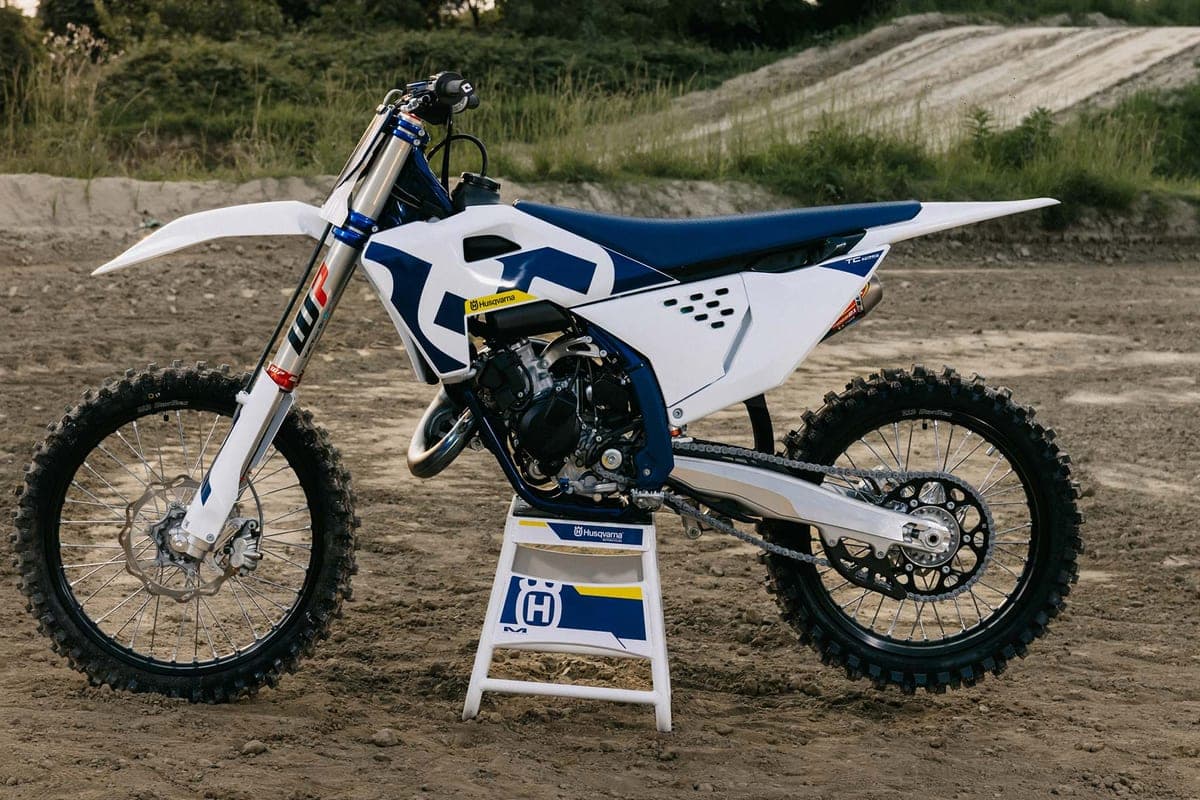 Husqvarna TC 125 parked on a track stand at a dirt track, right-side view highlighting the blue frame, white bodywork and yellow graphics
