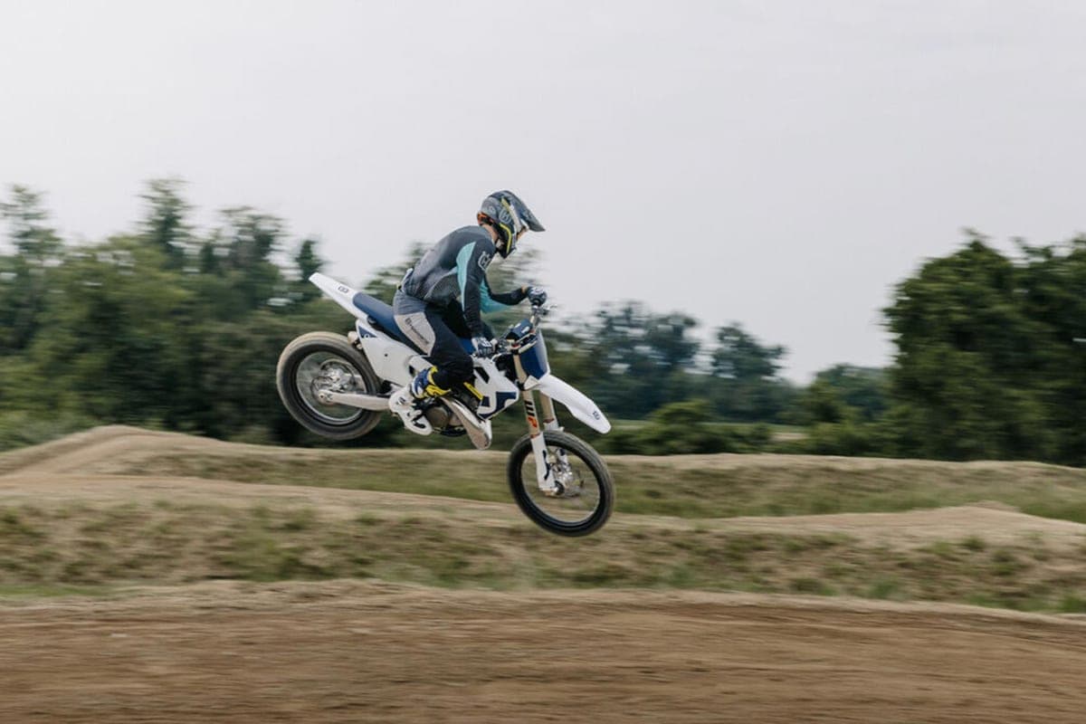 Rider in grey and teal gear catching air on a white Husqvarna TC 250 off a dirt jump, trees blurred behind from the panning shot