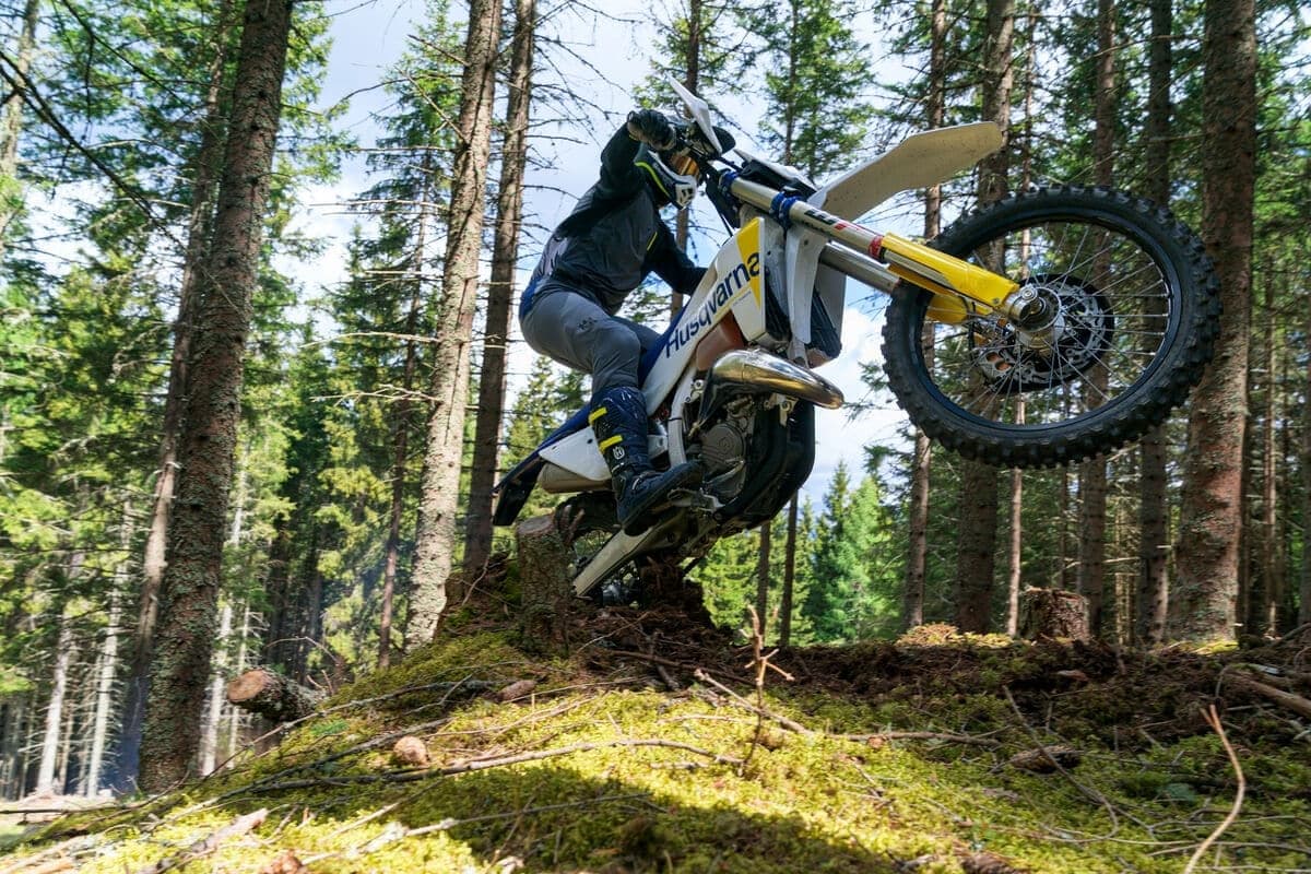 Rider in dark gear lofting the front wheel of a Husqvarna TE 150 over a mossy log in a pine forest trail