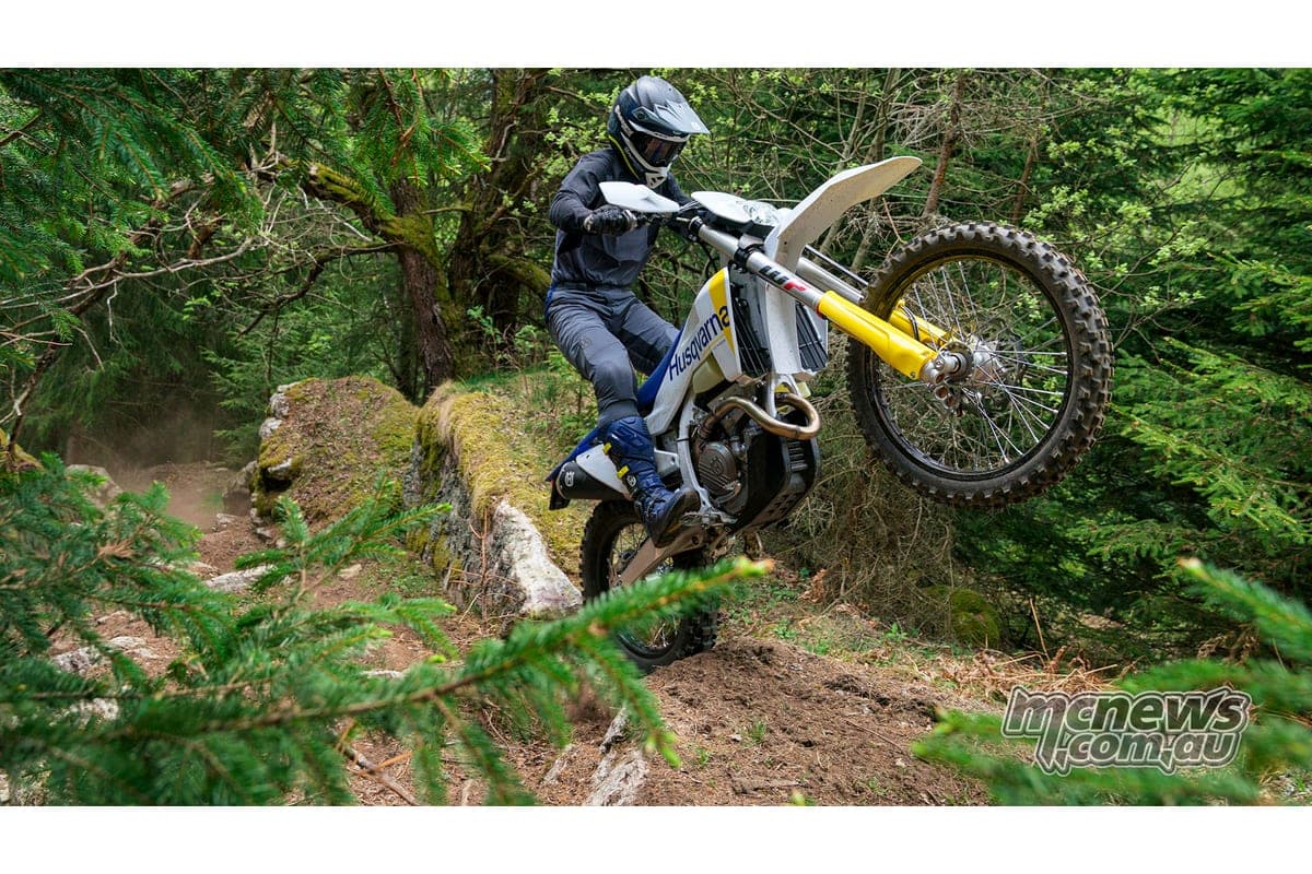 Rider in grey gear popping the front wheel of a Husqvarna TE 250 over a mossy log on a wooded trail, kicking up dust