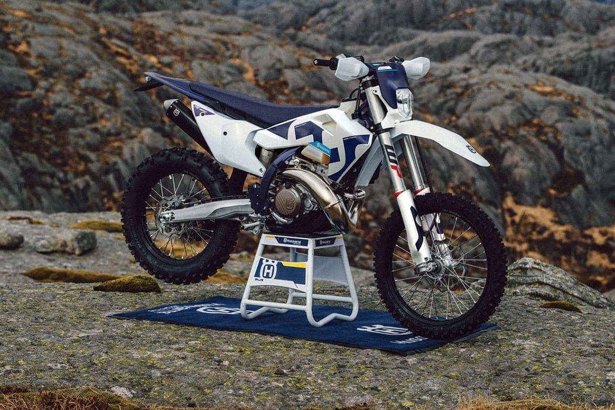 Husqvarna TE 300 in white with blue graphics, front three-quarter view on a rock stand outdoors, showing the two-stroke engine and spoked wheels