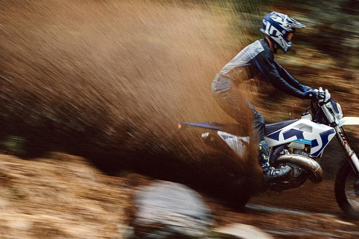 Rider in blue and grey gear leaning a Husqvarna TE 300 through a muddy corner, kicking up a heavy roost of wet dirt