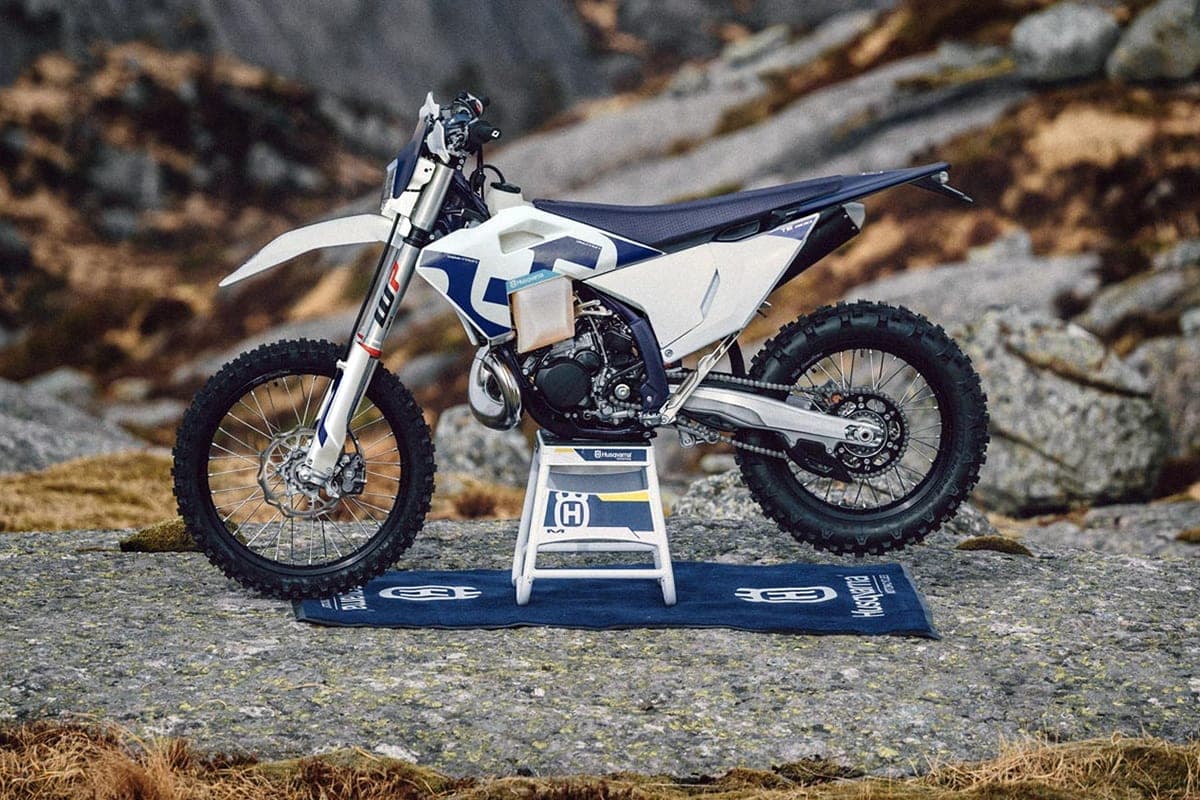 Husqvarna TE 300 rear three-quarter view parked on a workstand atop rocks, showing the chain, swingarm and knobby rear tire