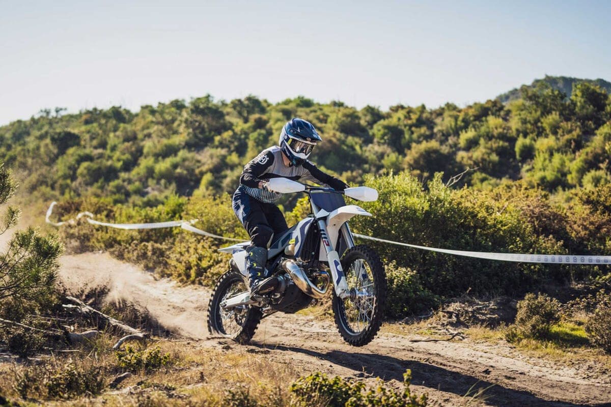 Husqvarna TX 300 rider in grey and black gear cresting a dusty trail bank, front wheel lifted with a roost of dirt behind