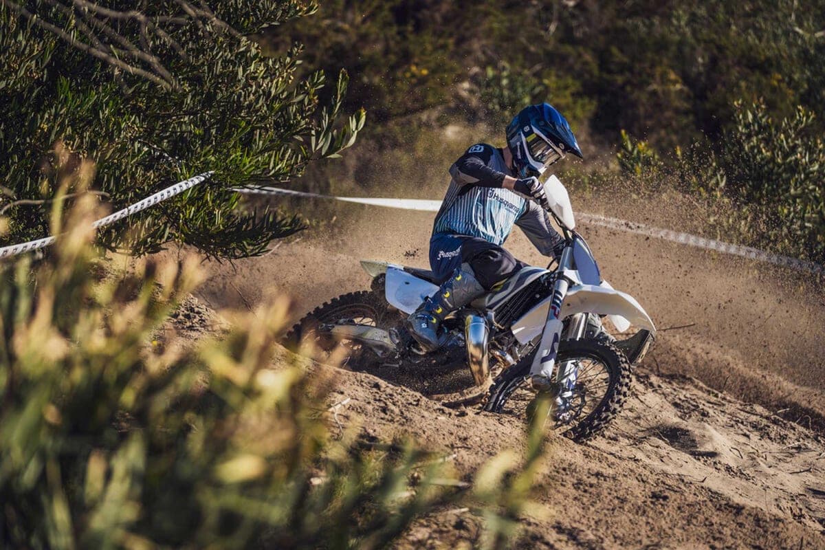 Husqvarna TX 300 rider leaning hard into a sandy corner, kicking up a wide spray of dust from the rear knobby tire