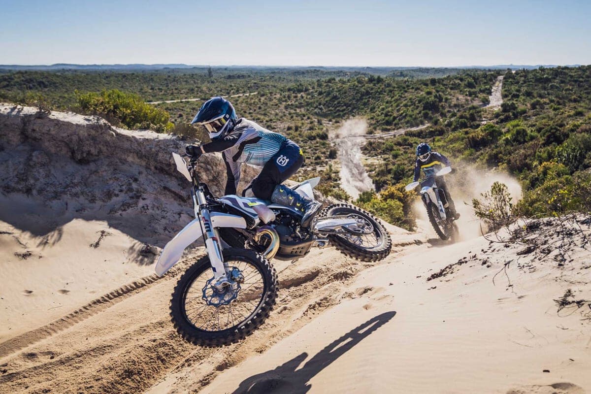 Two Husqvarna TX 300 riders descending a sandy dune trail, lead bike tipped into a turn with the second rider trailing dust behind