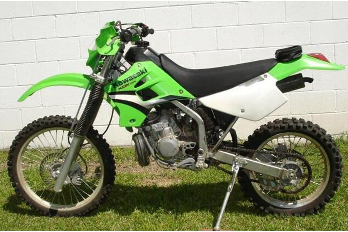 Kawasaki KDX200 parked on a kickstand against a cinderblock wall and grass, side view showing knobby tires, chain guard and rear taillight