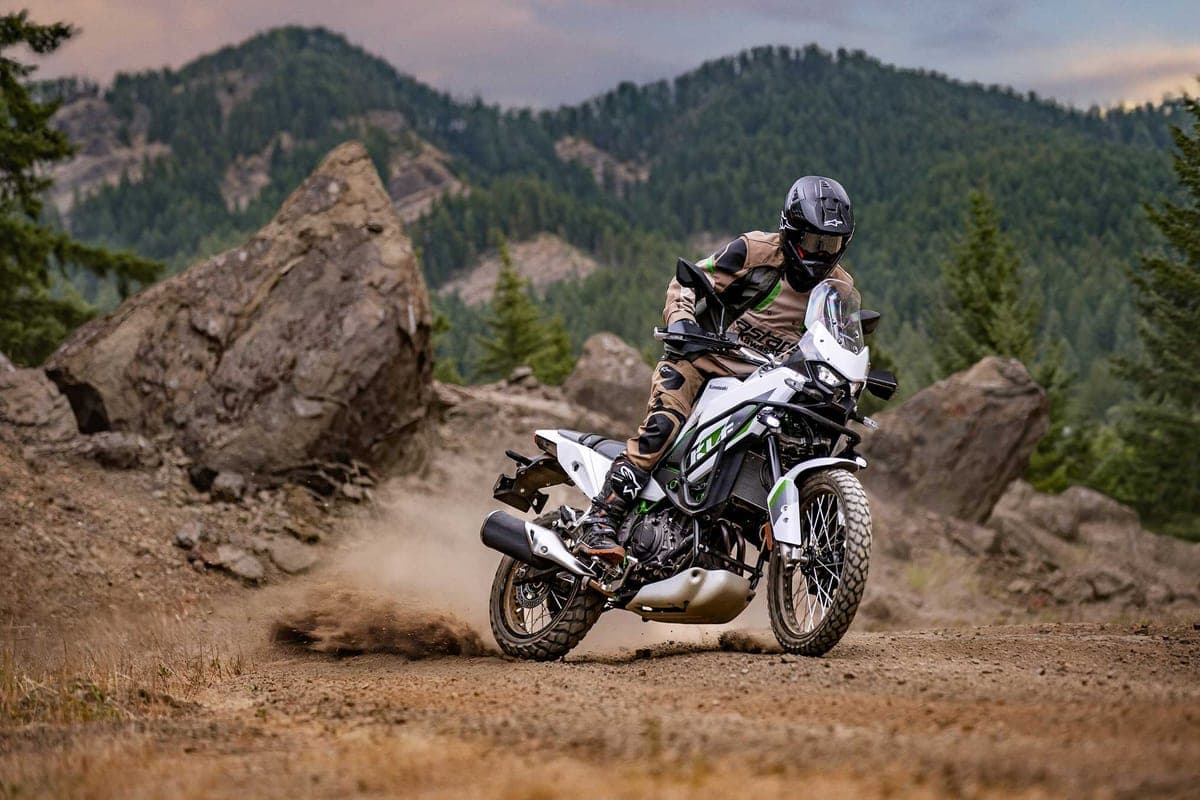 Rider in tan and black gear leaning the white KLE500 through a dusty rocky trail corner, kicking up a cloud of dirt behind the rear wheel