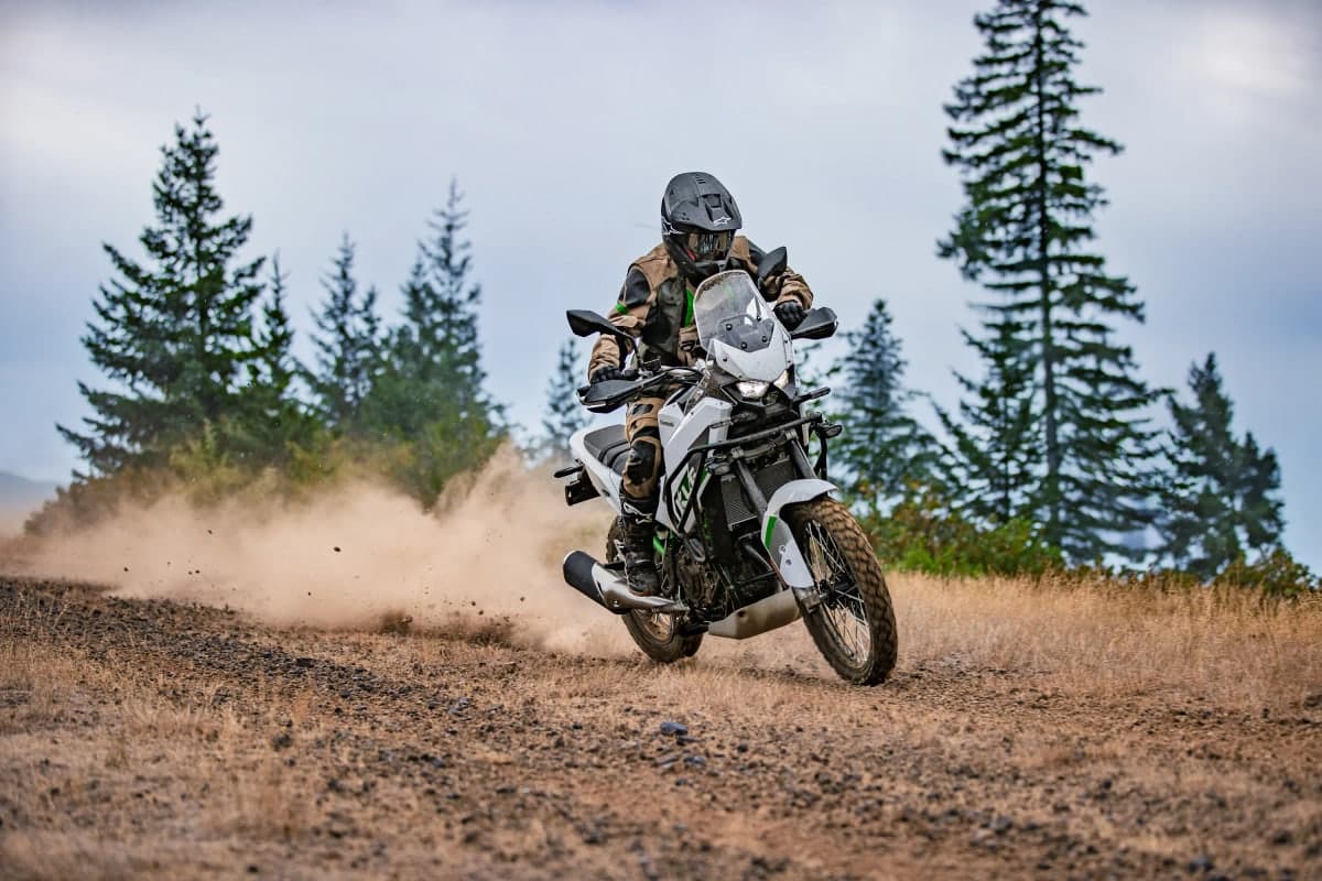 Rider in tan adventure gear kicking up dust on a white KLE500 SE on a forest dirt road