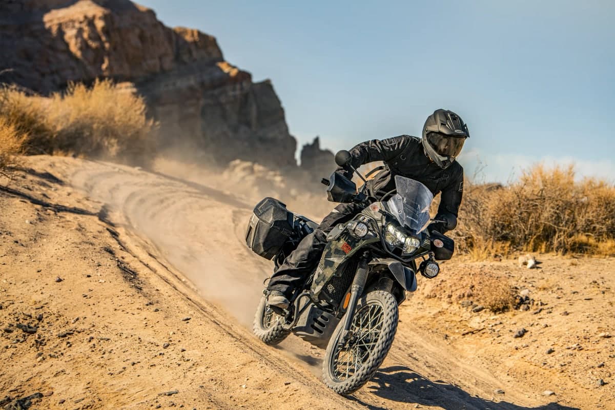 Rider on a camo KLR650 Adventure with saddlebags sliding through a dusty desert turn