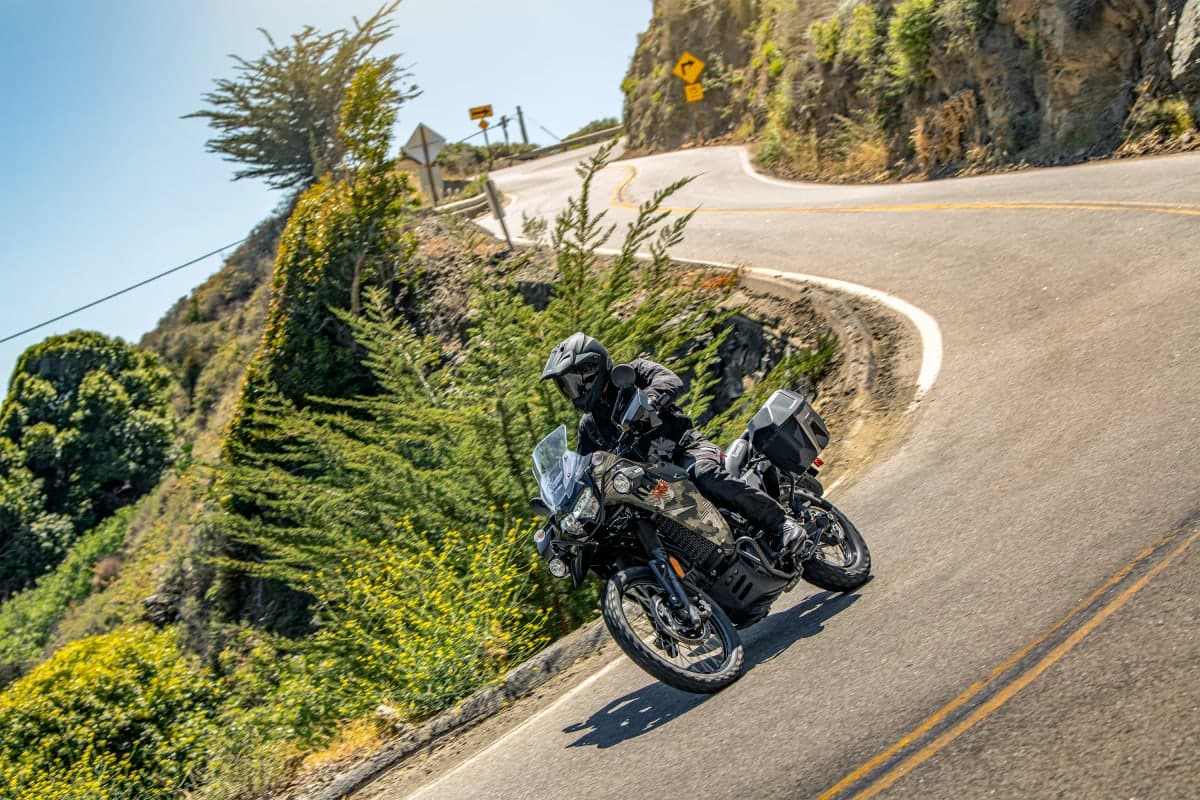 Rider leaning a camo KLR650 Adventure through a cliffside highway curve