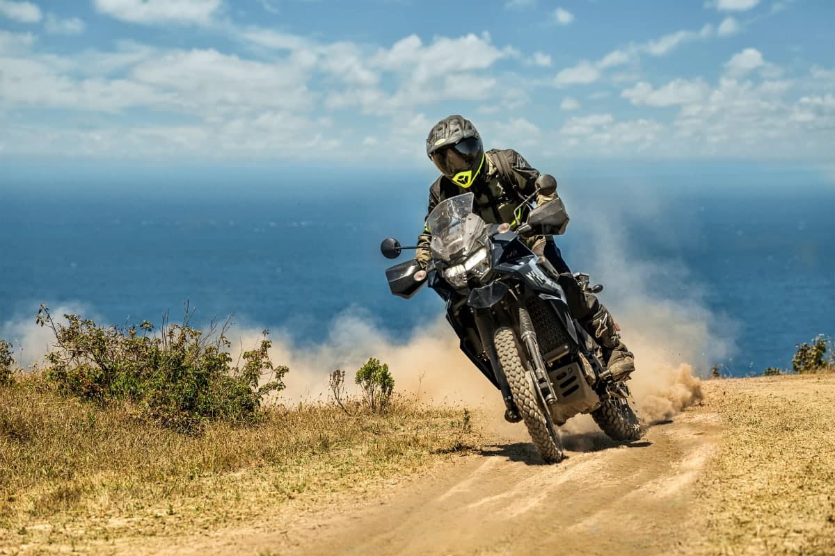 Rider sliding a KLR650 S through a dusty turn on a clifftop trail above the ocean