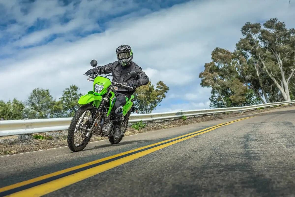 Rider in black gear riding a lime green Kawasaki KLX230 S ABS down a curving mountain road with guardrails and trees in the background