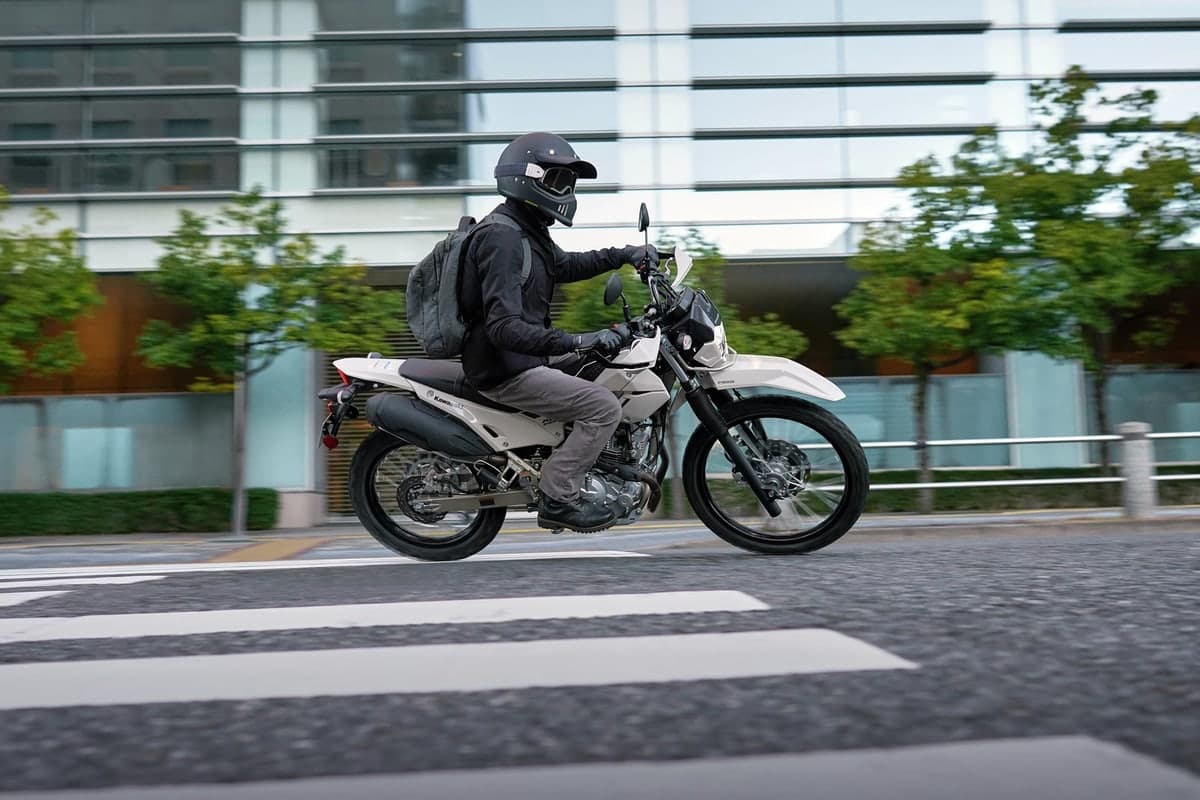 Rider in black gear and backpack riding a white KLX230 Sherpa past a glass office building, front wheel lofted slightly off the pavement