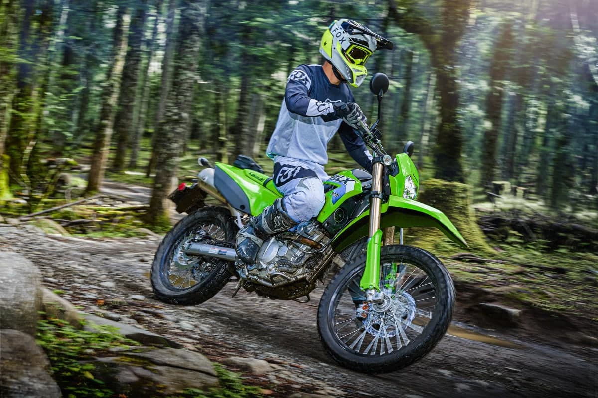 Rider in white and blue gear riding a green KLX300 over rocks on a forest trail