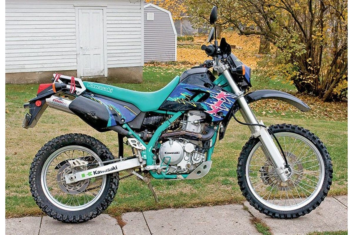 Kawasaki KLX650 in teal and black parked outdoors on a driveway, right-side view with aftermarket exhaust, knobby tires and autumn foliage in the background