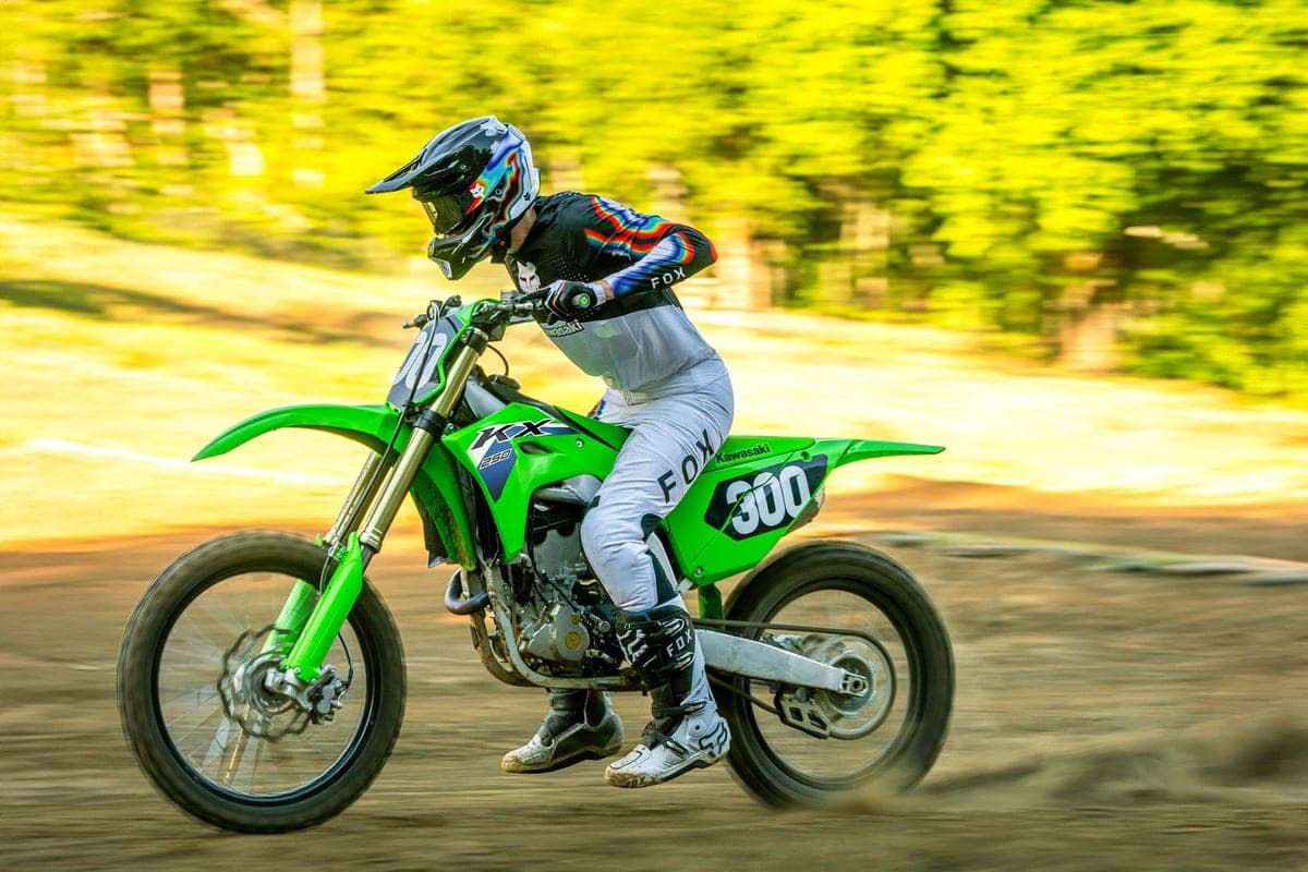 Rider in white and black Fox gear railing a green Kawasaki KX250F down a dirt track, front wheel light with motion blur trees behind