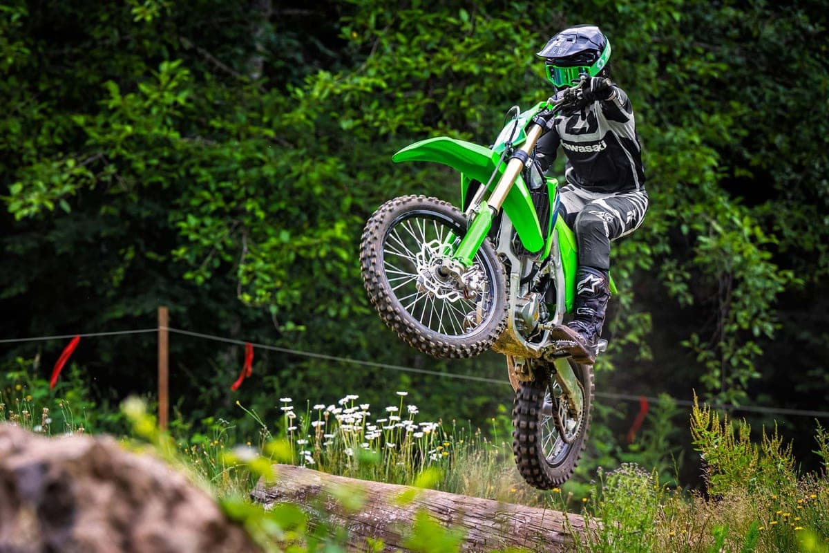 Rider in black and white gear catching air over a log on a green Kawasaki KX250X during a forest cross-country ride