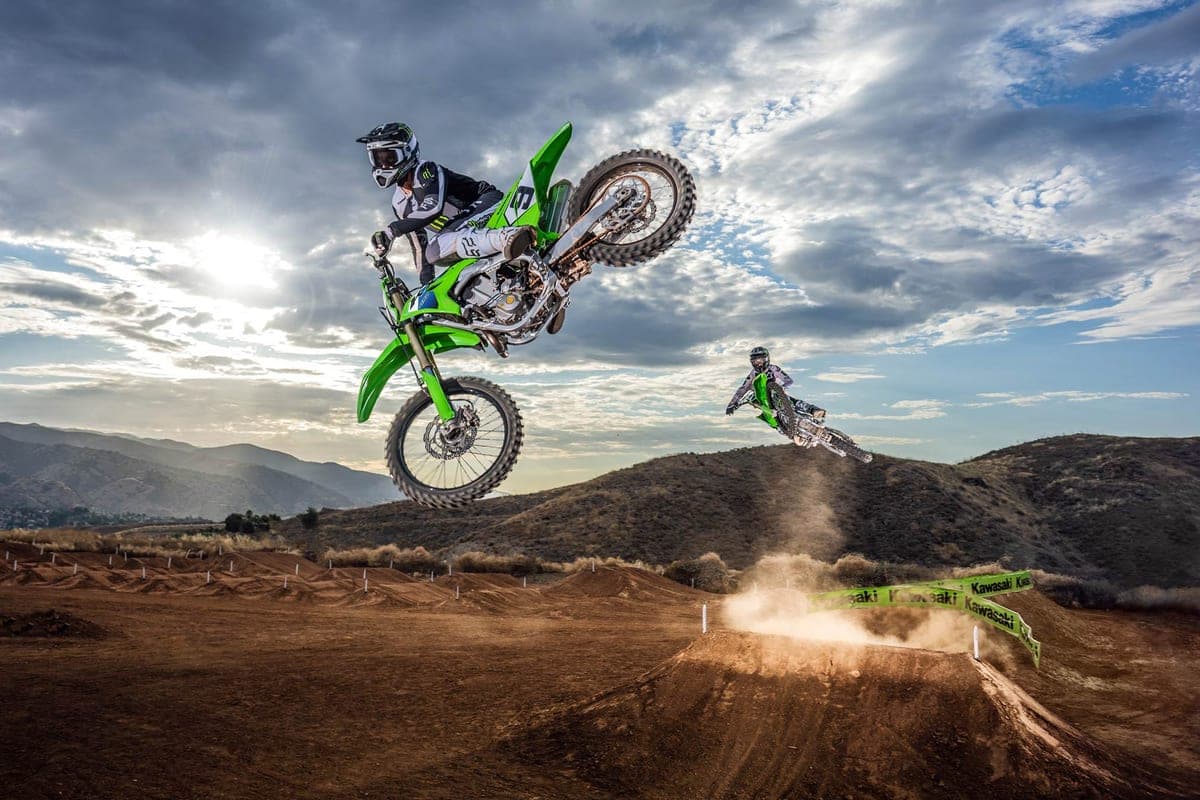 Rider in Monster Energy gear whipping a green Kawasaki KX450F sideways off a motocross jump, dust rolling off the landing behind