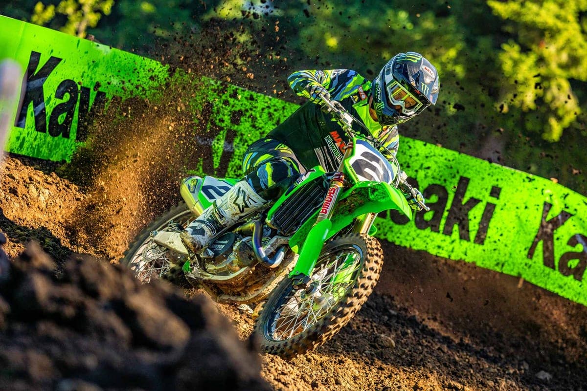Rider in green and black Monster Energy gear leaning a green Kawasaki KX450SR through a muddy corner, roosting brown dirt
