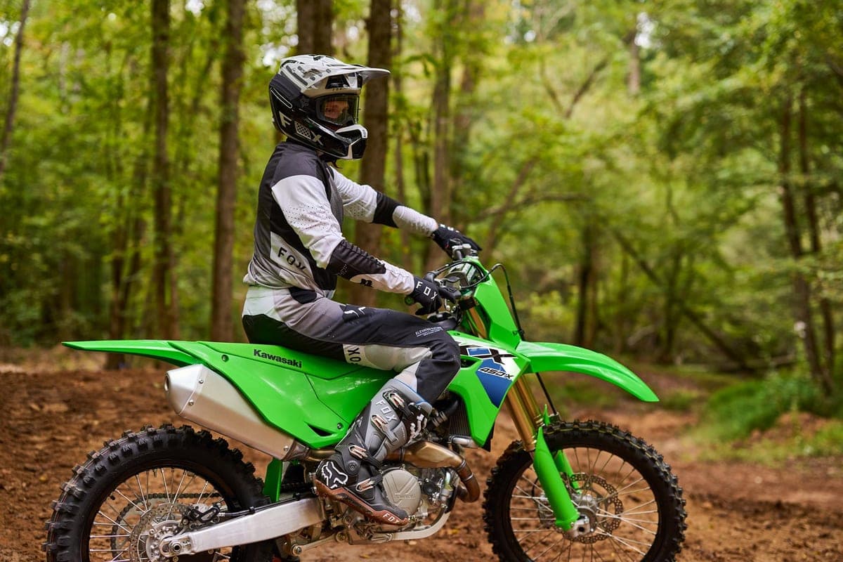 Kawasaki KX450X, rider in Fox gear riding it down a dirt trail through a green forest, front wheel lofted slightly over the rutted ground