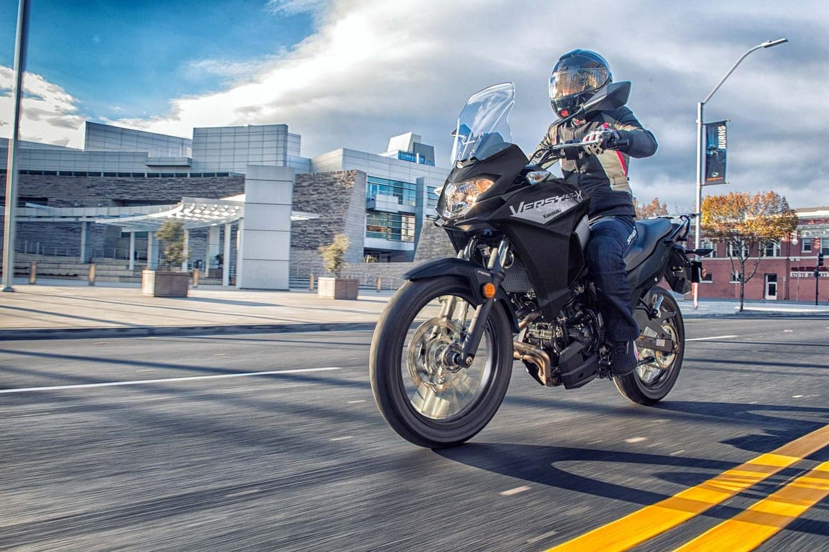 Rider in armored jacket and jeans wheelies a black Kawasaki Versys-X 300 through a city plaza past a modern glass building