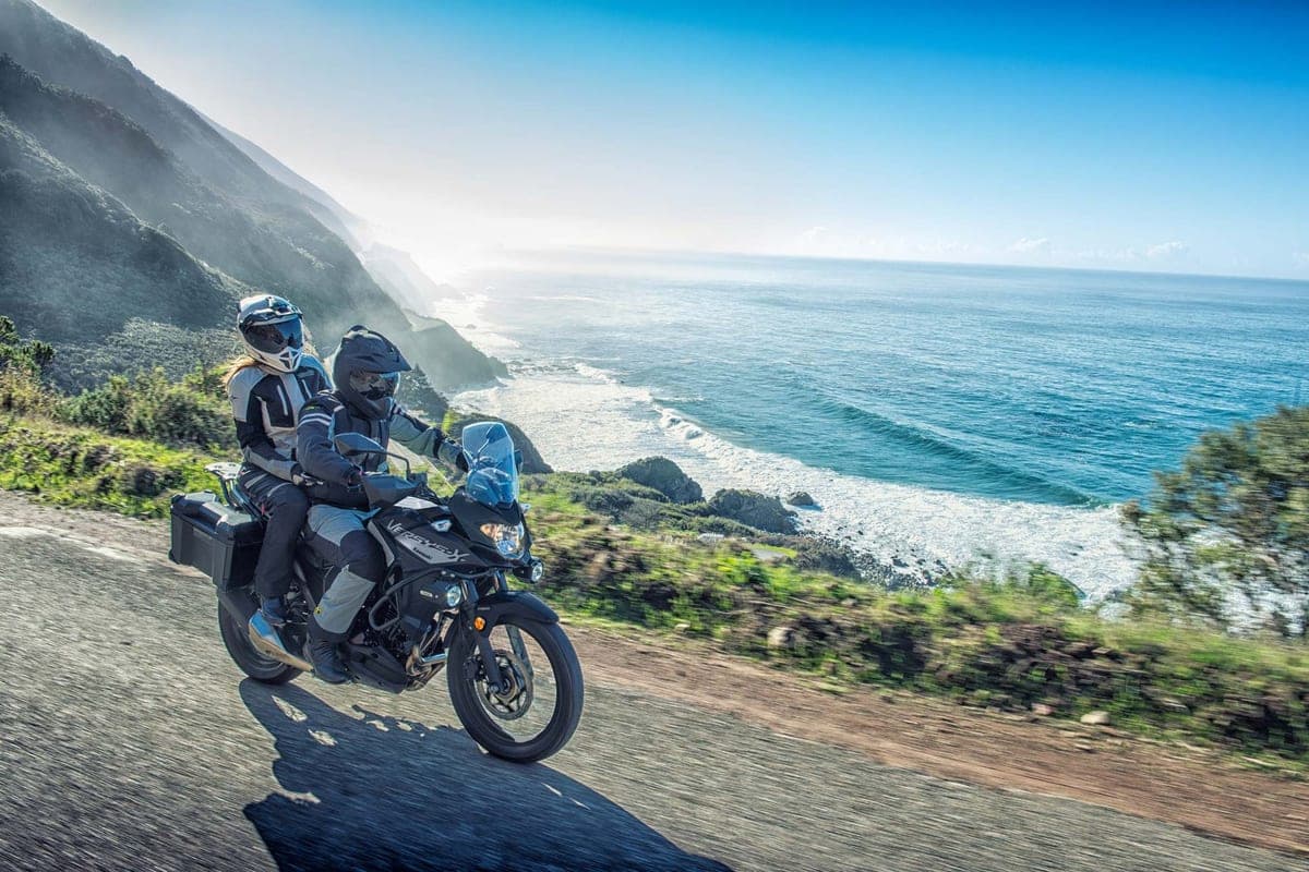 Two riders on a black Versys-X 300 with side cases lean into a coastal cliff road overlooking crashing ocean waves