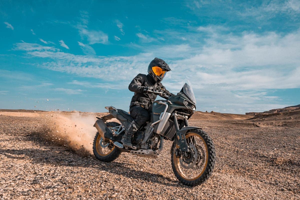 Rider in black gear leaning the Kove 800X Pro through a gravel desert turn, kicking up a spray of dust under a bright blue sky