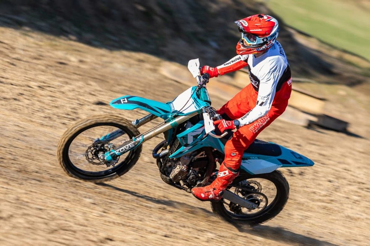Rider in red-and-white gear leaning into a berm on a teal-and-white Kove MX250, kicking up a spray of dirt on a motocross track