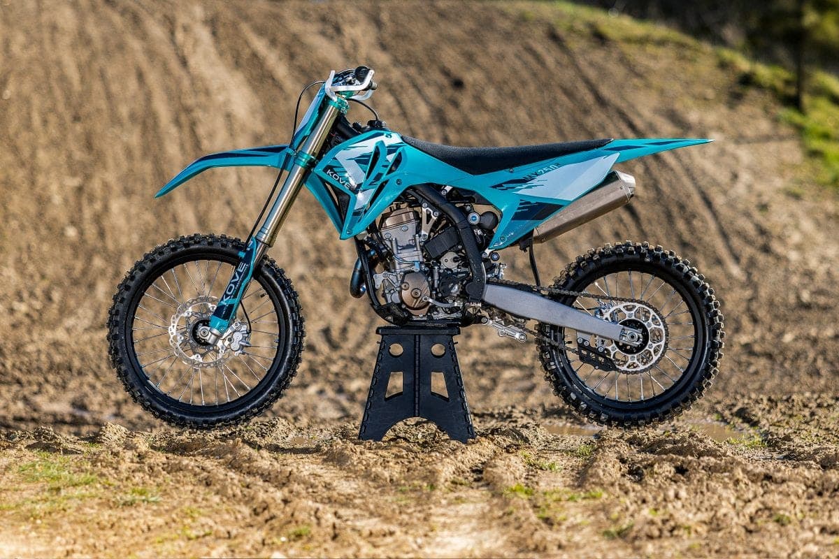 Kove MX250 in teal and light blue livery, left-side view on a bike stand on a motocross track with a dirt berm behind it