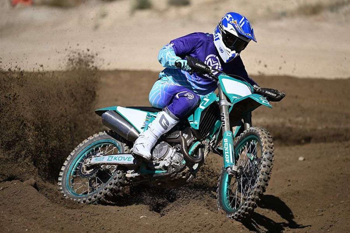 Rider in purple and teal gear leaning into a berm on a teal Kove MX450, dirt spraying from the rear knobby tire on a motocross track